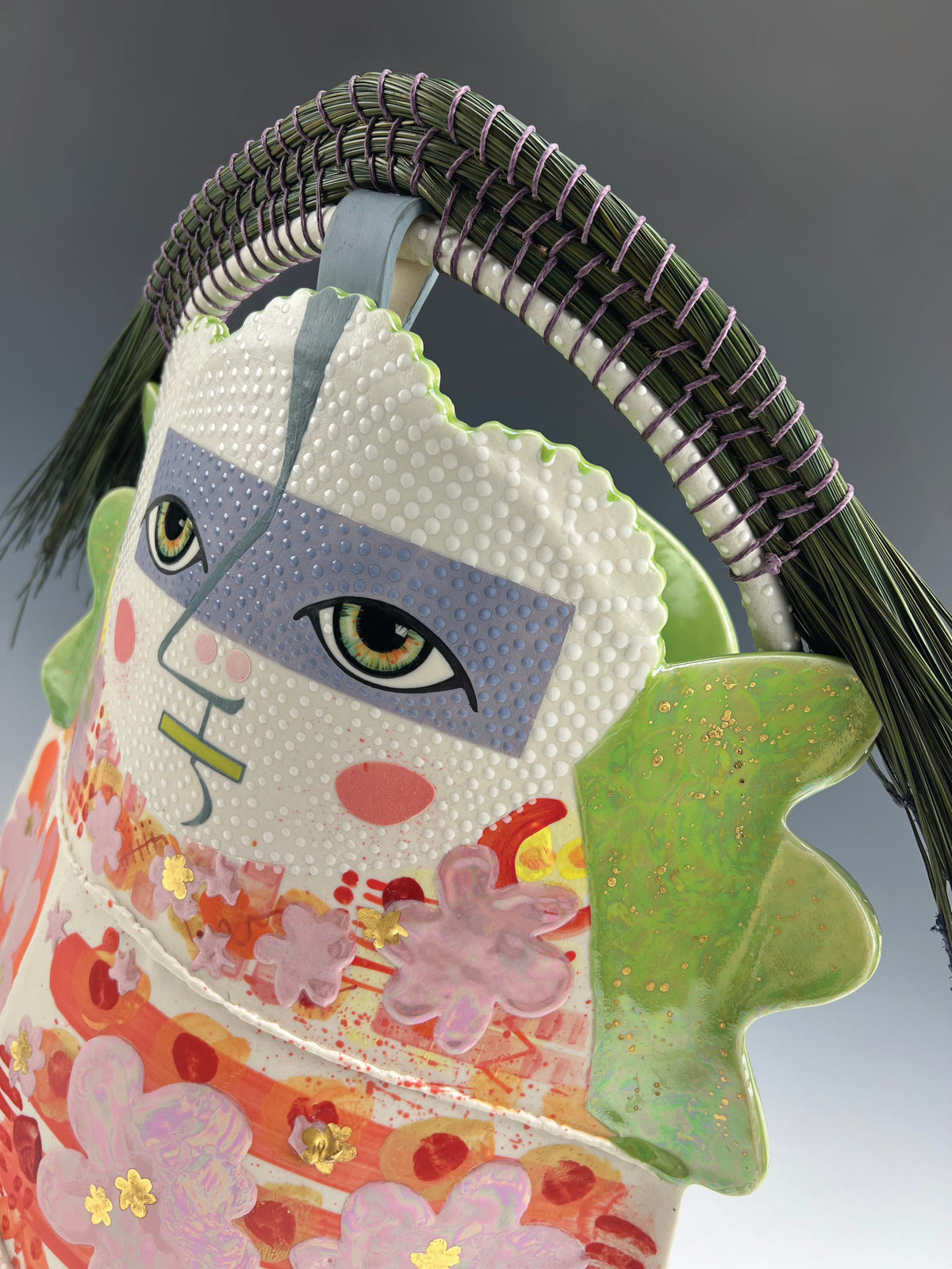 Artist: Jennifer Rosseter; A close-up of a ceramic sculpture featuring a stylized face with intense green eyes and crown of green Florida Long Leaf pine needles. The body is primarily orange, red, pink, and green.