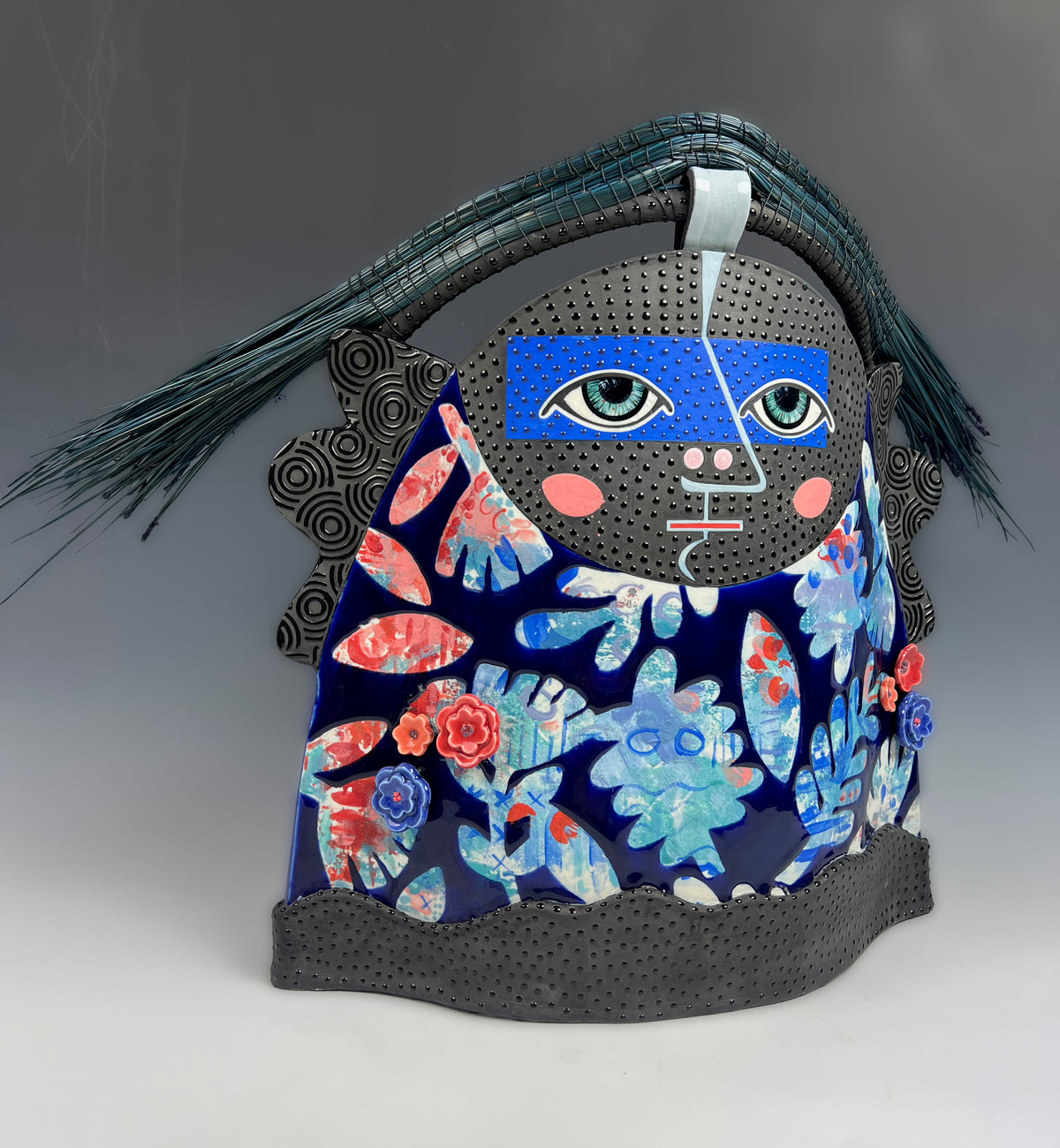 Artist: Jennifer Rosseter; Sculptural ceramic art, featuring a stylized face with a crown of blue-green Florida Long Leaf pine needles overhead. The body is designed with a colorful floral and leaf patterns in red, pink, and light blue. The piece has graphic black accents and a black base.